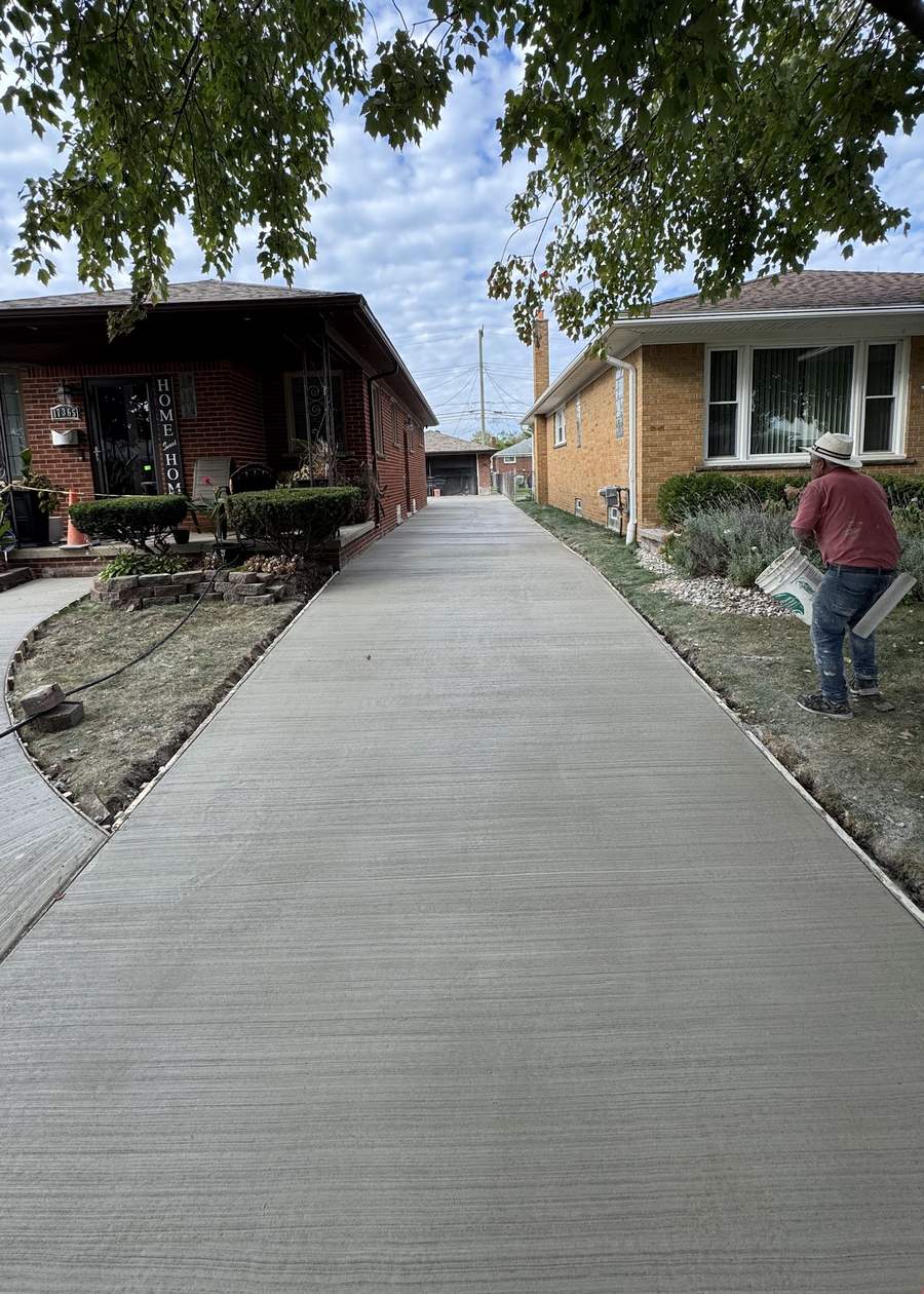 A freshly poured broom-finish concrete driveway between two brick houses in Metro Detroit, a Cabrera crew member finishing the edge