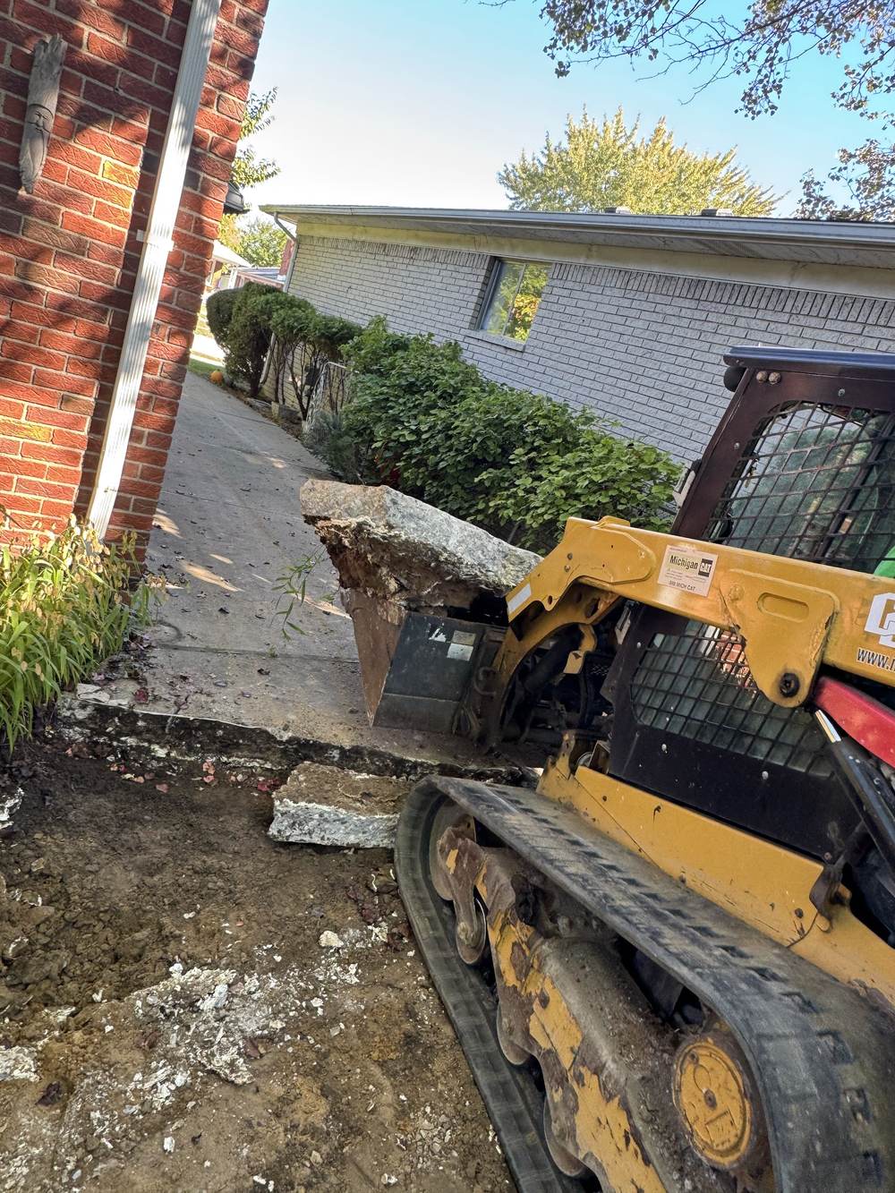 A Cat skid steer lifting out a slab of old concrete driveway beside a red brick house